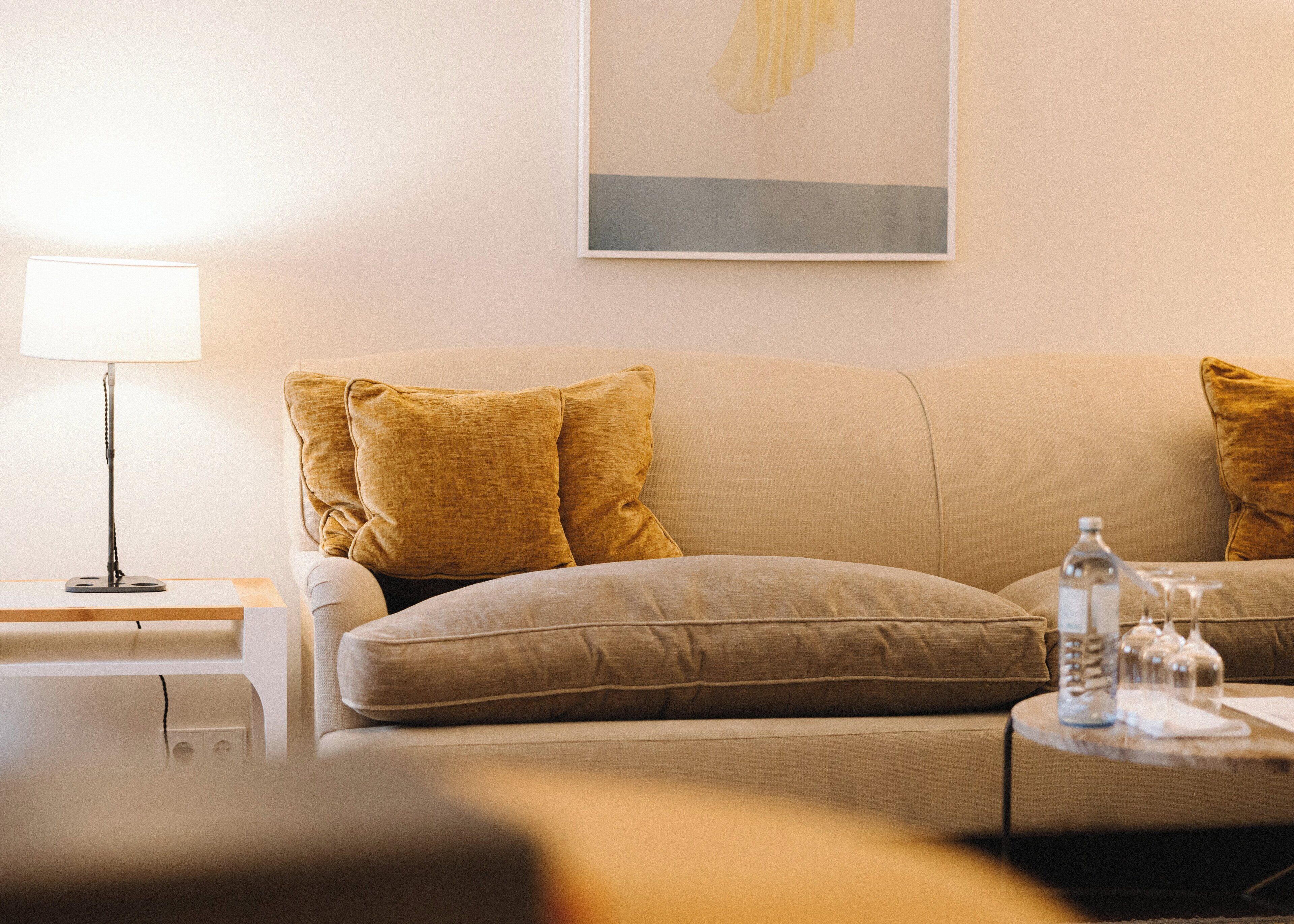 Una sala de estar con un sofá color beige, dos cojines amarillo mostaza, una mesita de noche con una lámpara y una pequeña mesa con una botella y vasos.