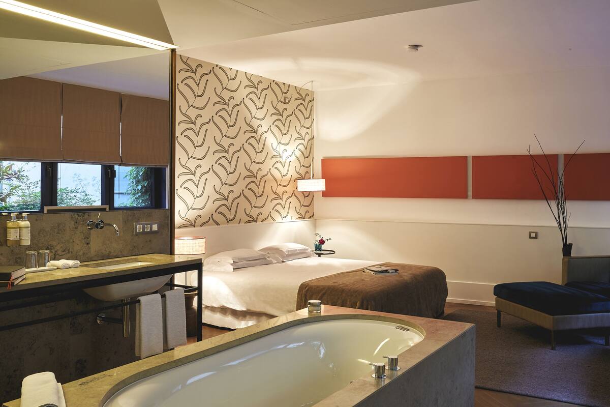A modern hotel room with a large bathtub, a separate bathroom with a sink and mirror, a bed with a headboard, and a small bedside table.