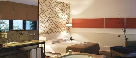 A modern hotel room with a large bathtub, a separate bathroom with a sink and mirror, a bed with a headboard, and a small bedside table.