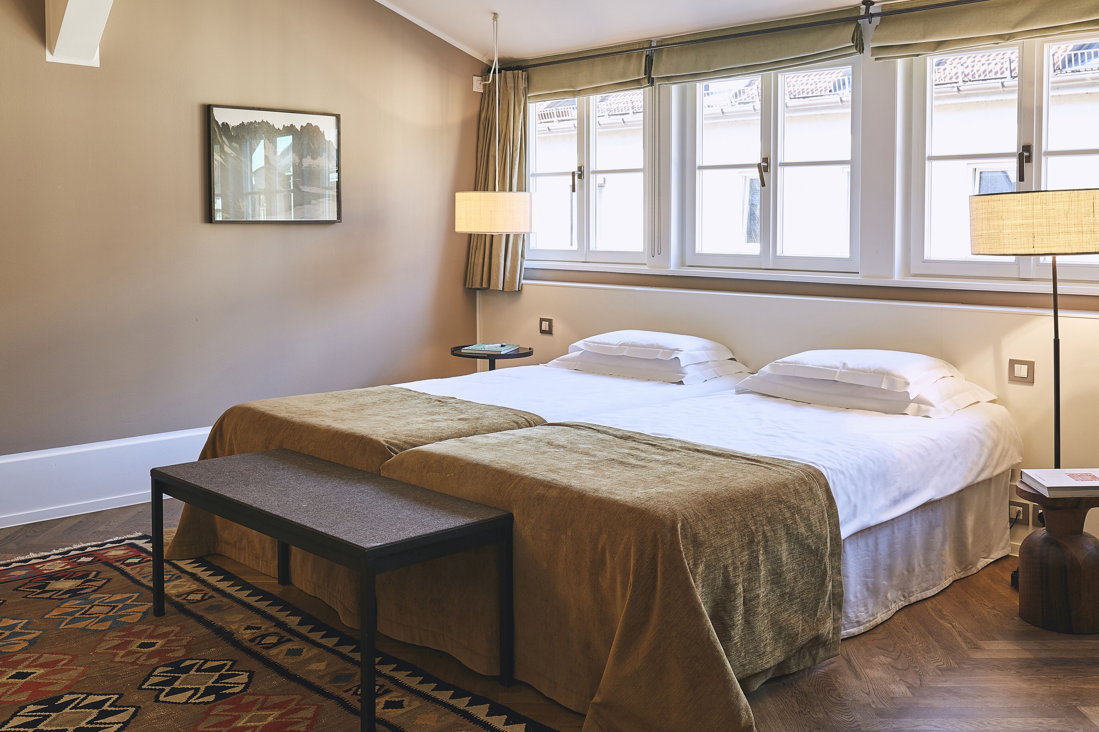 Una habitación de hotel con cama, mesitas de noche, un banco, un tapete y una ventana con cortinas.