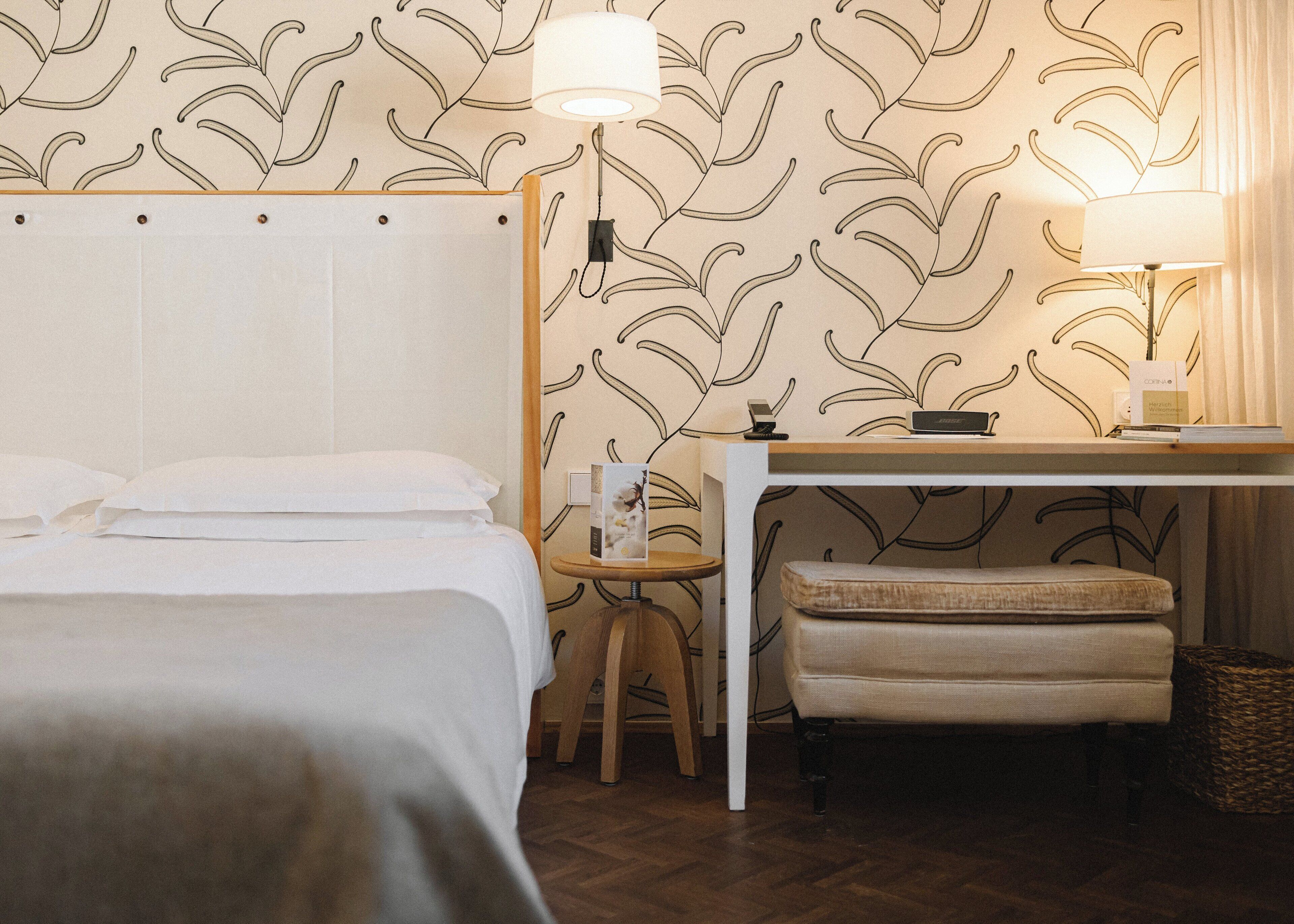 A hotel room with a bed, a desk, a chair, a lamp, and a small table with a book and a phone.