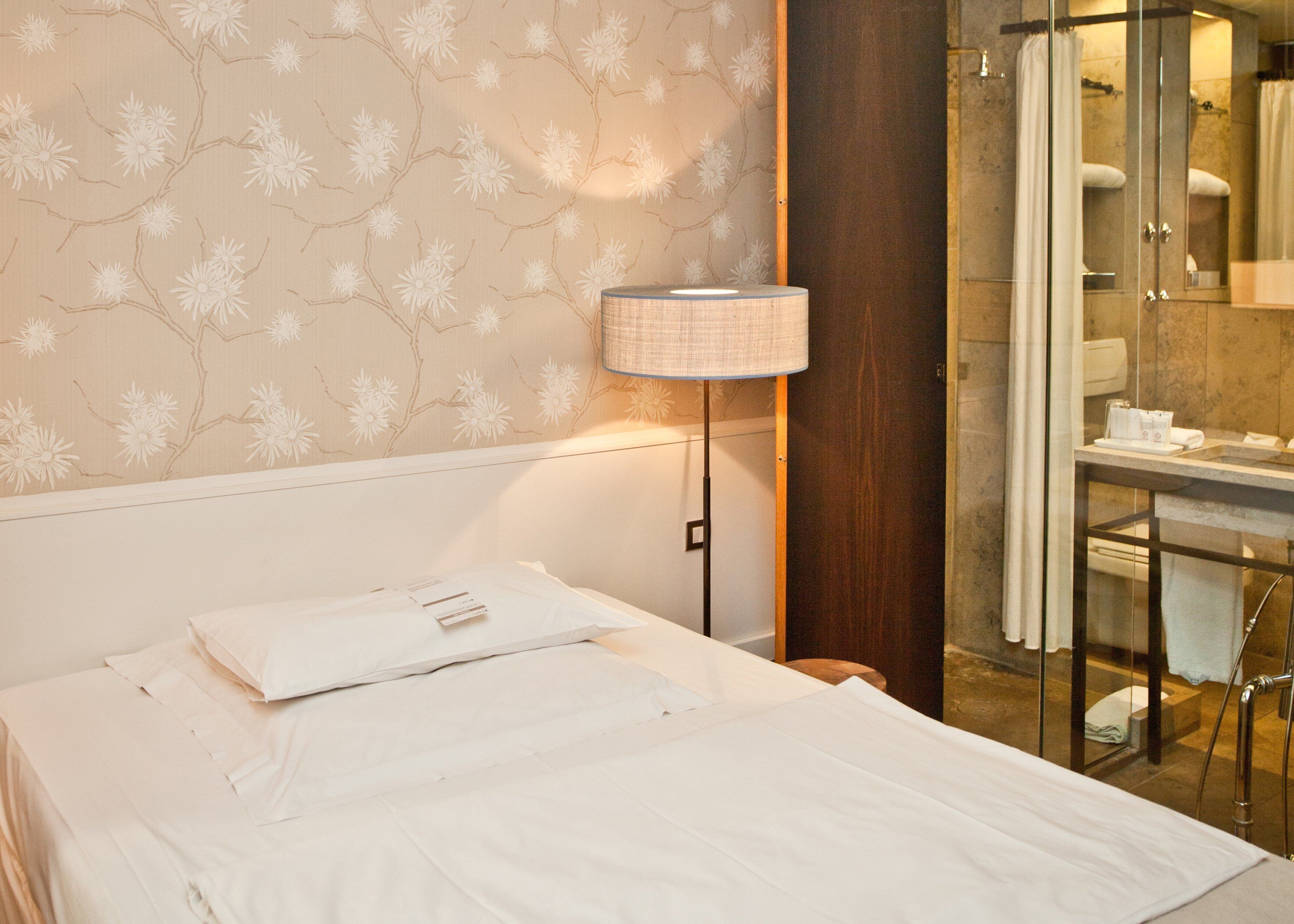 Quarto de hotel com cama, mesa de cabeceira, luminária e área de chuveiro.