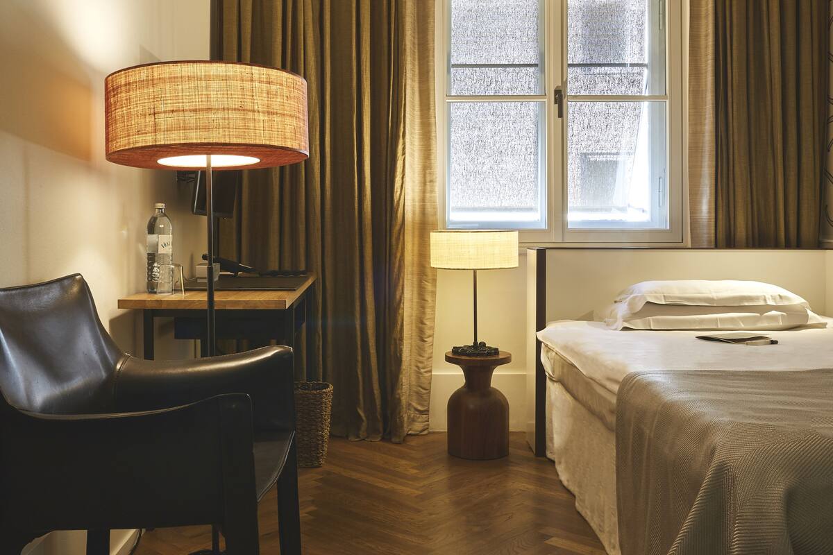 A hotel room with a bed, a desk, a chair, a lamp, and a window with curtains.