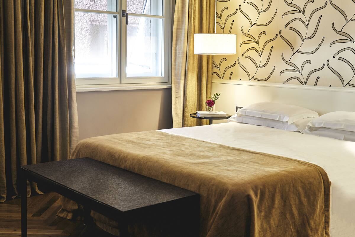 A hotel room with a large bed, a bedside table, a lamp, and a view of the outdoors through a window.