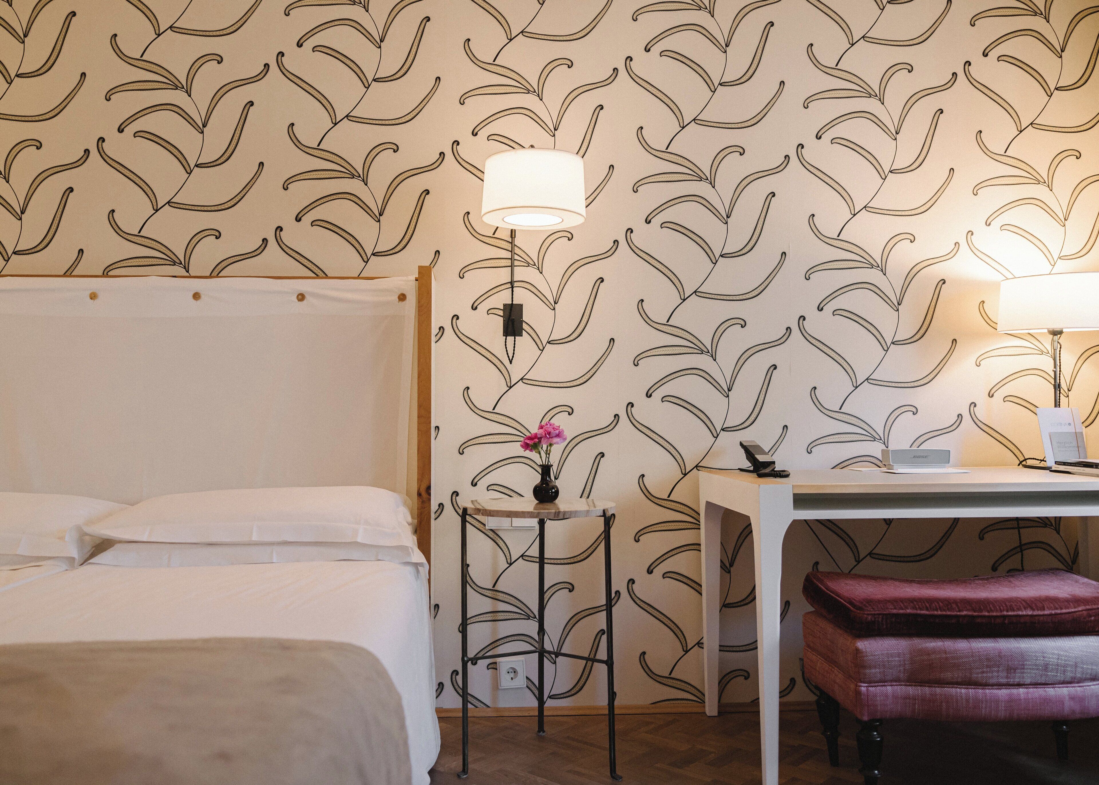 A hotel room with a bed, a desk with a lamp, a small table with a vase of flowers, and a red bench.