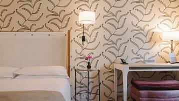A hotel room with a bed, a desk with a lamp, a small table with a vase of flowers, and a red bench.