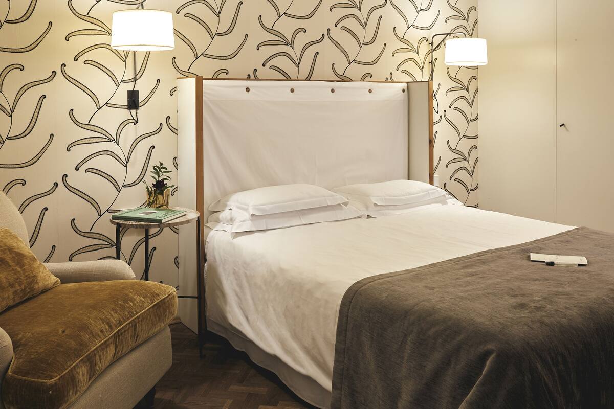 A neatly made bed with white linens, a brown upholstered armchair, and a small table with a lamp.