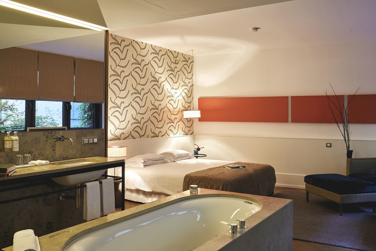 Une chambre d’hôtel moderne dotée d’une grande baignoire, d’un espace salle de bain séparé, d’un lit avec une tête de lit et d’une petite table de chevet.