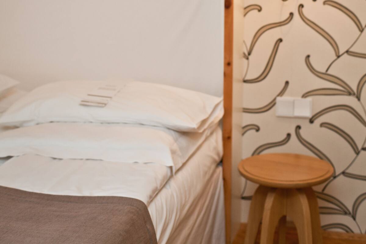 A bed with white linens, a wooden nightstand, and a wall-mounted lamp.