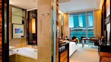 A modern hotel room with a large bed, a desk, a chair, a TV, and a bathroom with a bathtub and a large mirror.