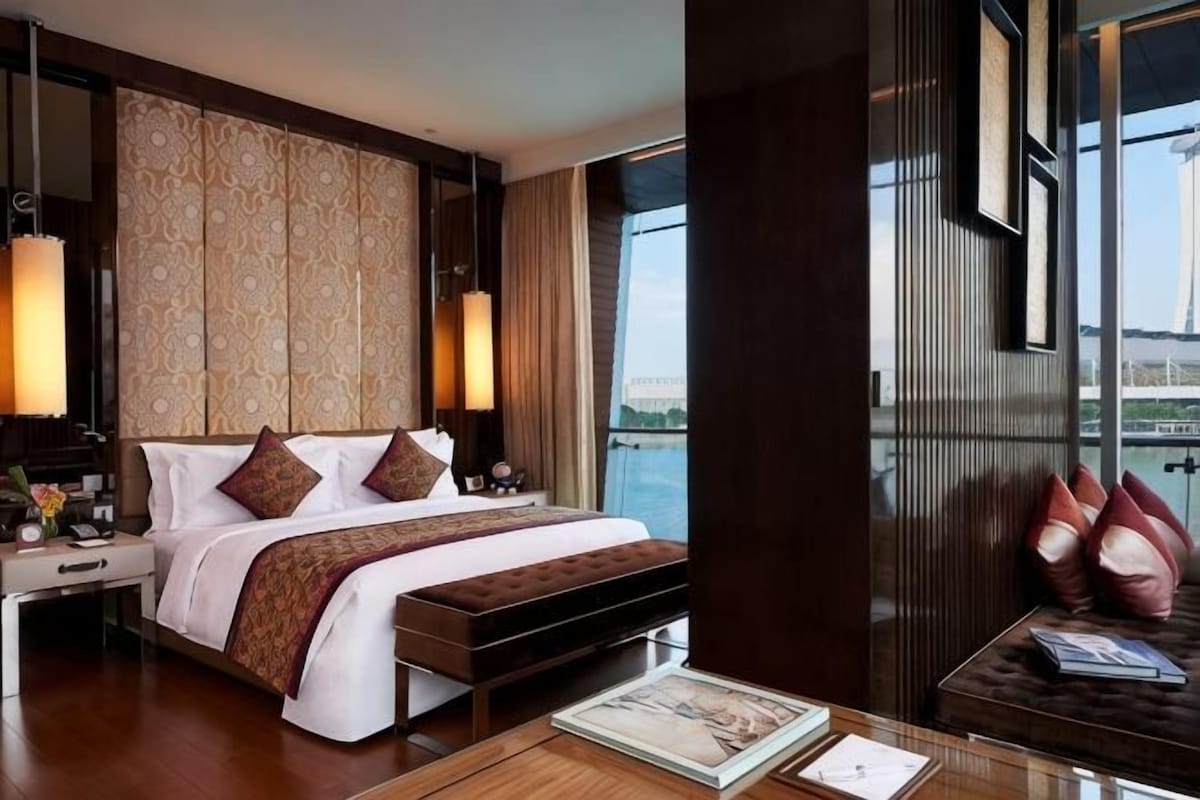 Habitación de hotel con una cama grande, un banco de madera, una mesita de noche y vistas al agua.