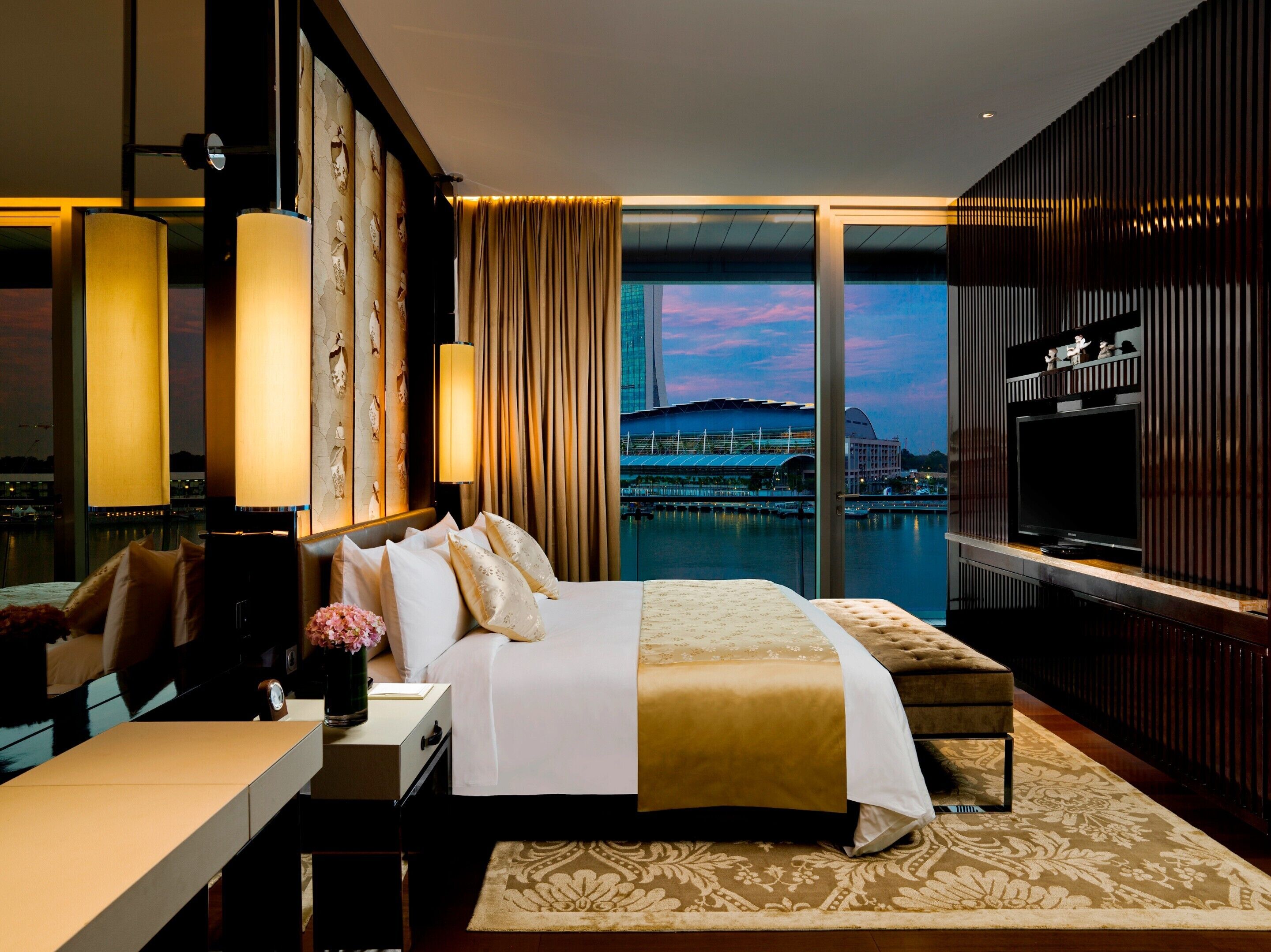 Habitación de hotel con una cama grande, escritorio, televisión y vistas a un paisaje urbano y al agua.
