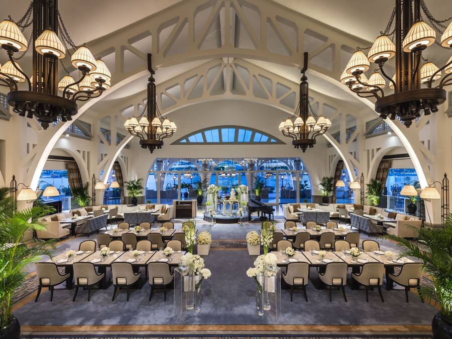 A spacious hotel lobby with high ceilings, chandeliers, and a central stage. Tables are set with white flowers and the lobby has large windows.