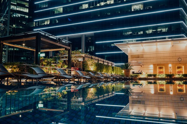 Zona de piscina moderna de hotel con sillones, piscina de cristal y un edificio con líneas iluminadas.