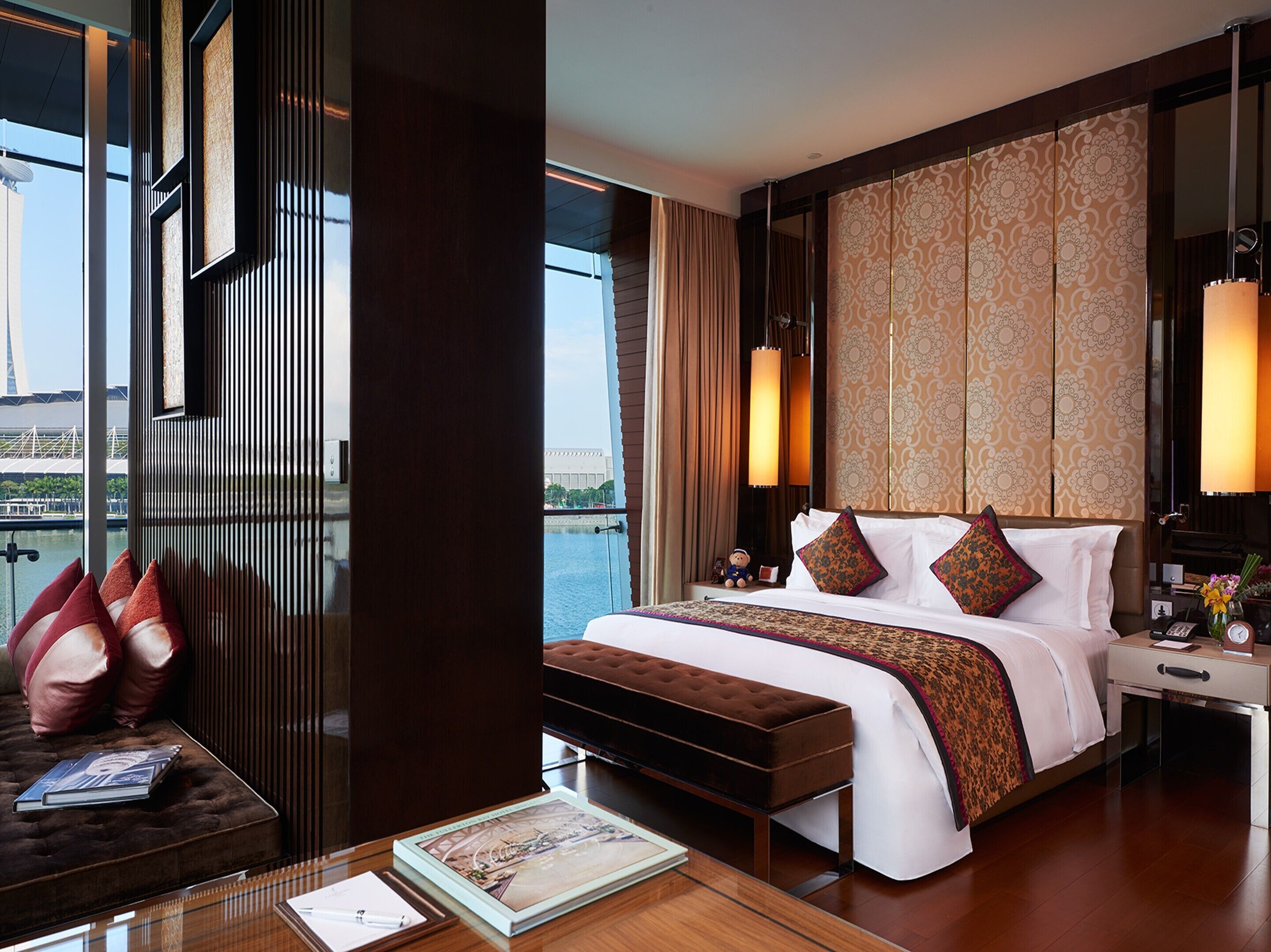 A hotel room with a large bed, a sitting area with a sofa and a table, a view of a bridge and water outside, and a decorative wall panel.