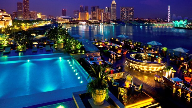 A rooftop pool area with lounge chairs and umbrellas, overlooking a city skyline at night.