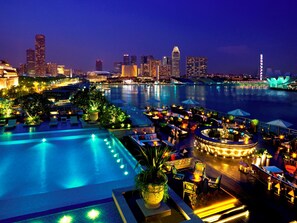 Zona de piscina en la azotea con sillones reclinables y sombrillas, con vistas al horizonte de la ciudad por la noche.