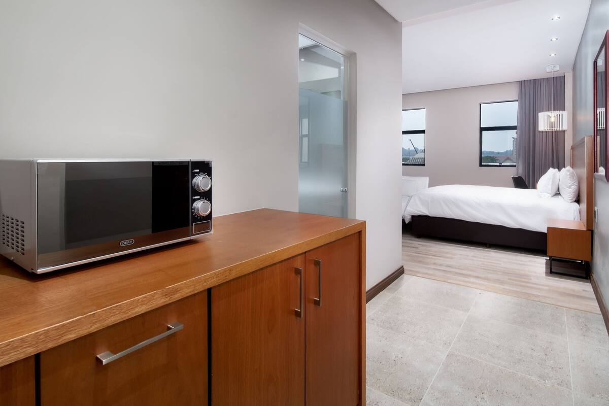 Une chambre d’hôtel moderne avec un four à micro-ondes posé sur un comptoir en bois, un lit avec du linge de maison blanc, une table de chevet et une vue sur la ville à travers de grandes fenêtres.