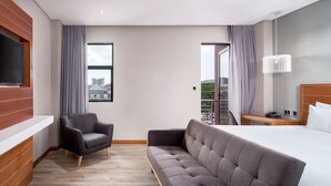 A modern hotel room with a large bed, a grey sofa, a black armchair, and a TV mounted on a wooden panel.