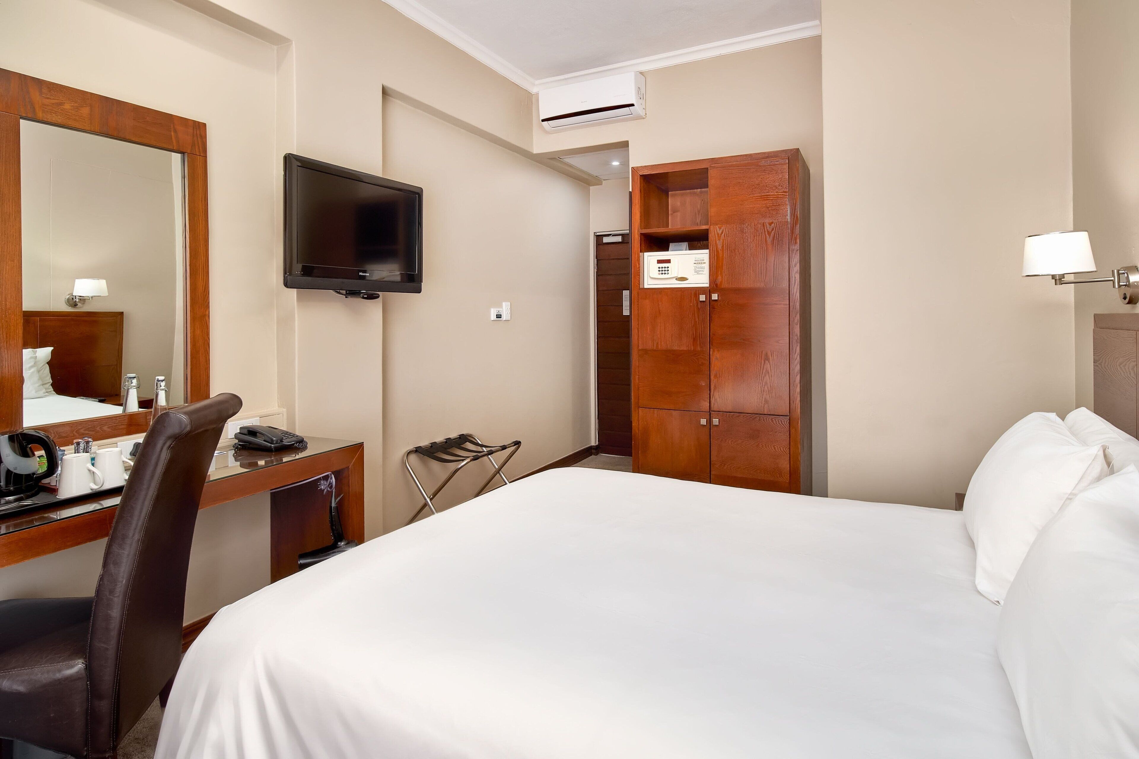 A hotel room with a bed, a desk with a chair, a television, a wardrobe, and a lamp.