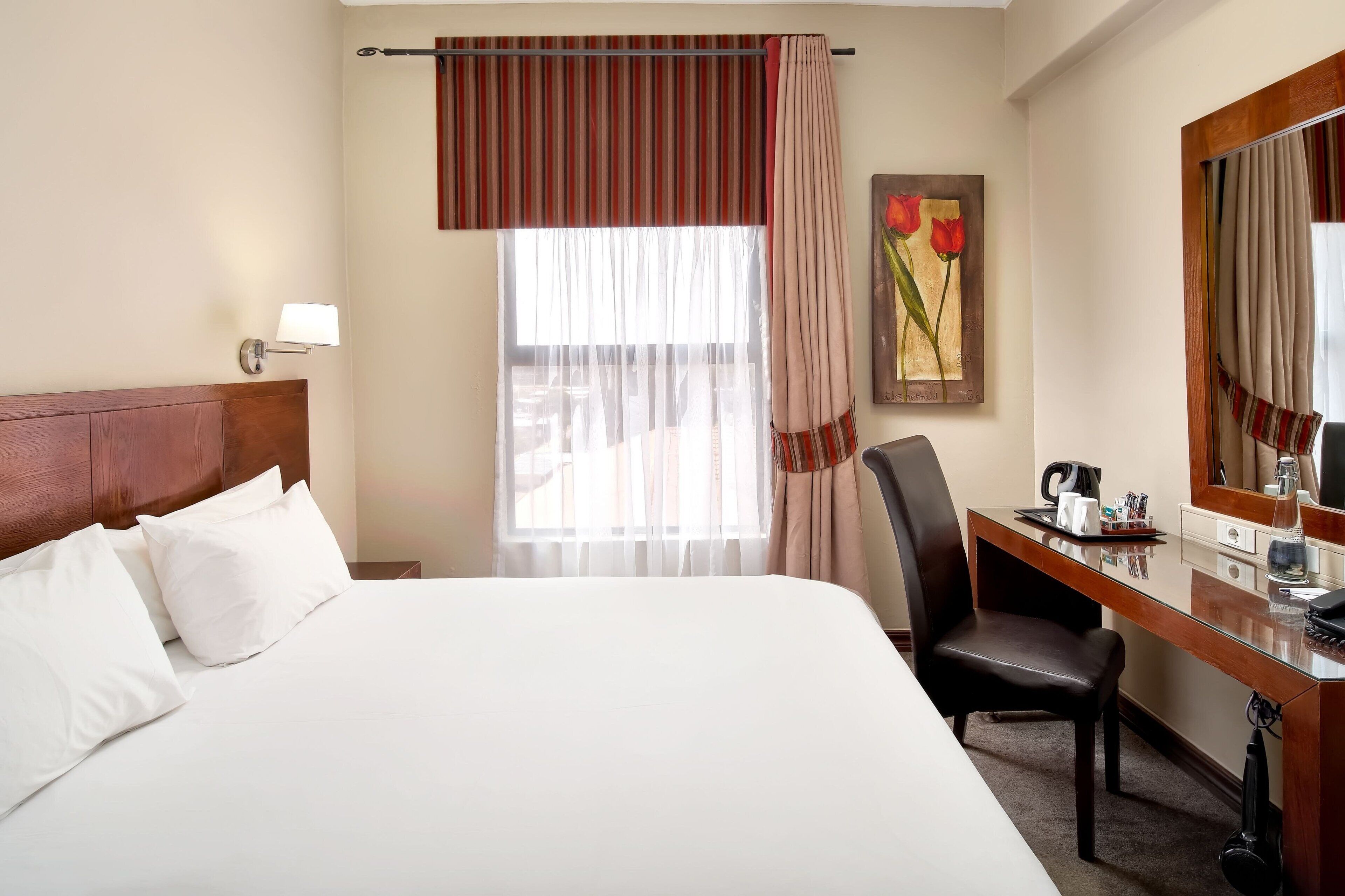 A hotel room with a large bed, a desk, a chair, a lamp, and a painting on the wall.