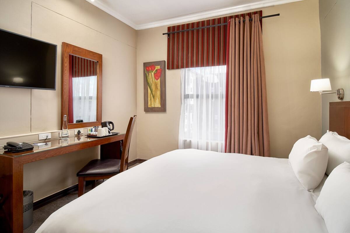 Une chambre d’hôtel comprenant un lit, un bureau avec un téléphone, une chaise, un téléviseur et une fenêtre avec des rideaux.