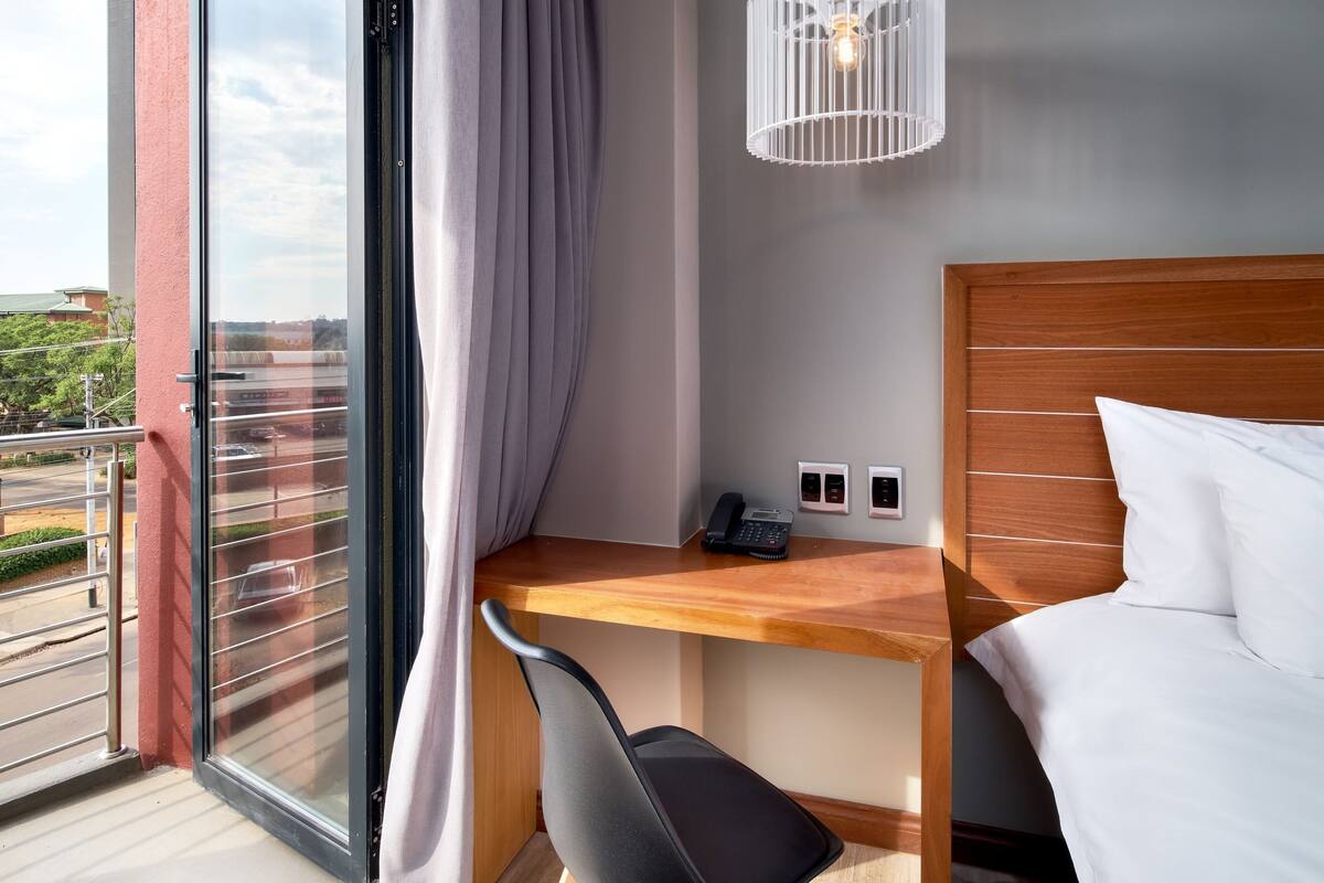 Une chambre d’hôtel avec un lit, un bureau doté d’un téléphone et d’une lampe, une chaise, et une vue sur l’extérieur.