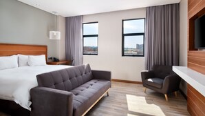 A modern hotel room with a large bed, a sofa, a chair, a TV, and two windows with curtains.
