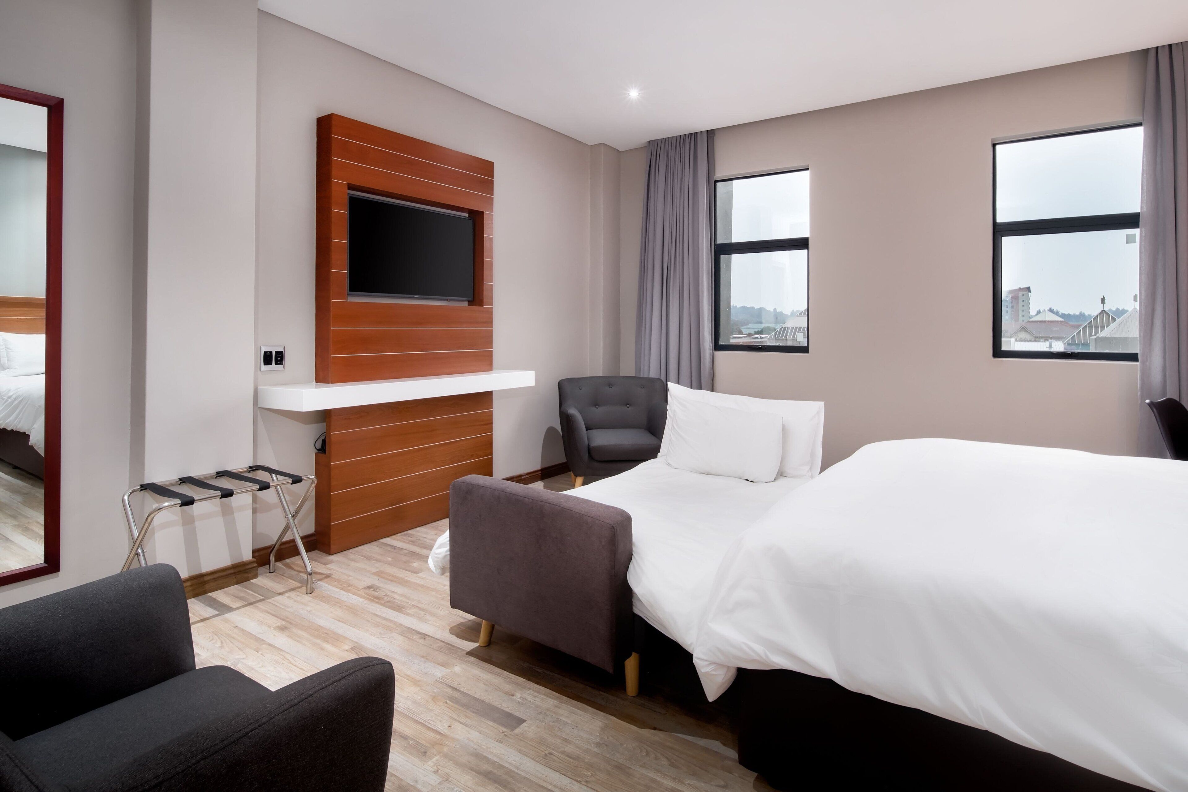 Ett hotellrum med en säng, en tv som är monterad på en träpanel, en stol och ett fönster med utsikt över staden.