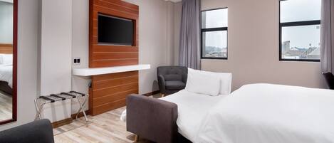Ett hotellrum med en säng, en tv som är monterad på en träpanel, en stol och ett fönster med utsikt över staden.