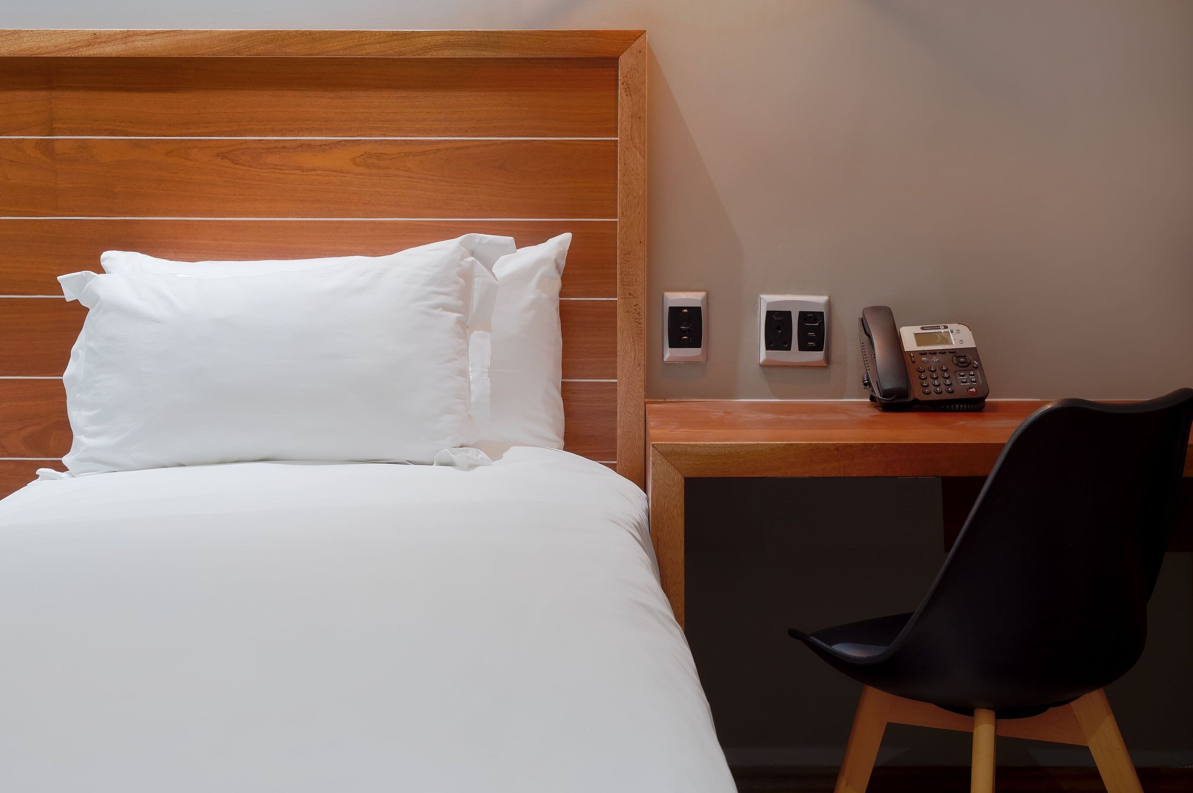 A hotel room with a wooden headboard, a bed with white linens, a desk with a telephone, and a black chair.
