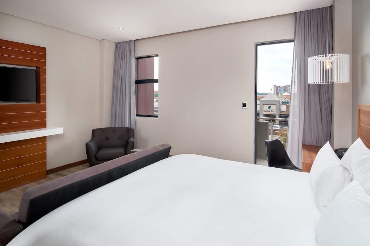 Une chambre d’hôtel dotée d’un grand lit, d’un téléviseur fixé sur un panneau en bois, d’un coin salon avec un fauteuil et d’une vue sur la ville par la fenêtre.