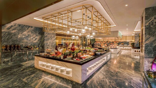 A luxurious hotel lobby with a marble reception desk, a modern chandelier, and a well-stocked buffet area.