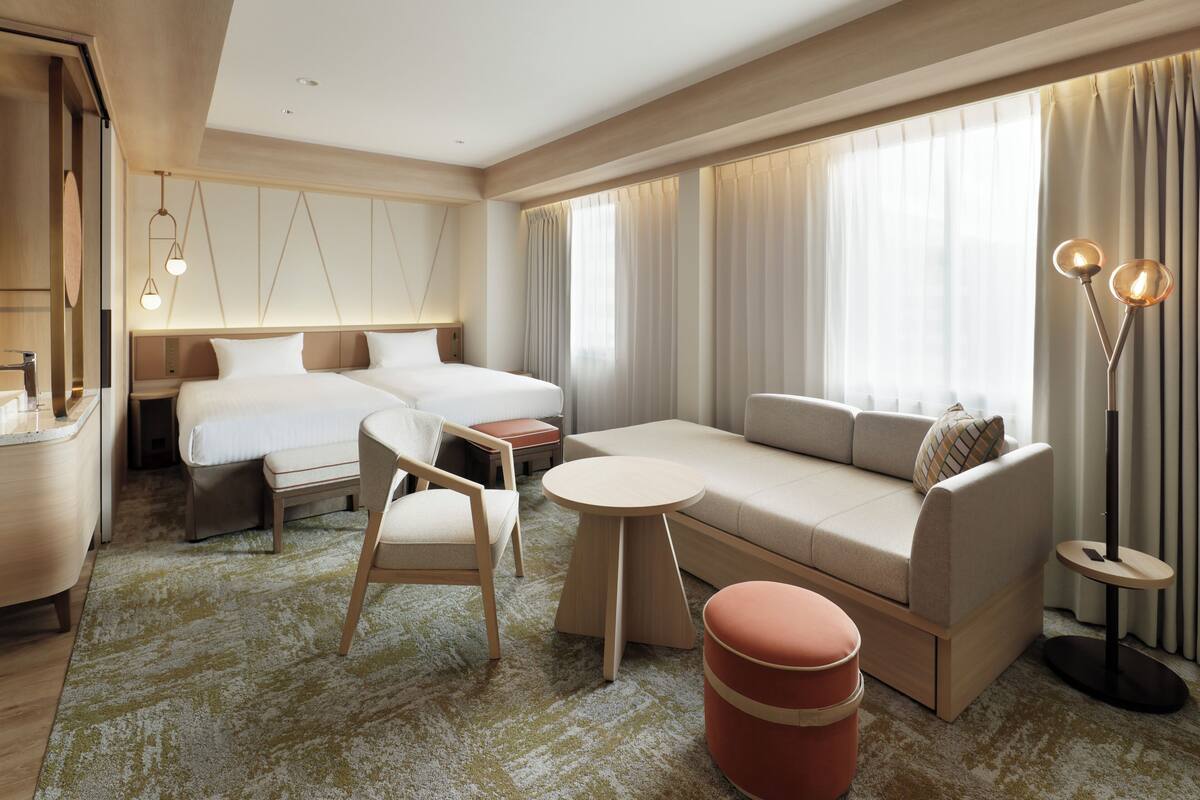 Quarto de hotel moderno com cama, sofá, poltrona, mesa redonda pequena e abajur.