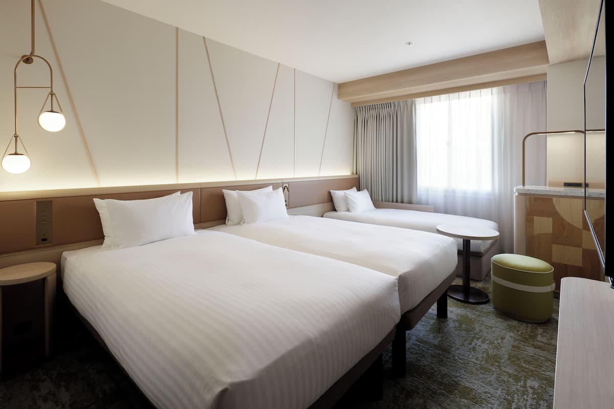 Quarto de hotel com duas camas, uma mesa redonda pequena, um sofá, um espelho e uma janela com cortinas.
