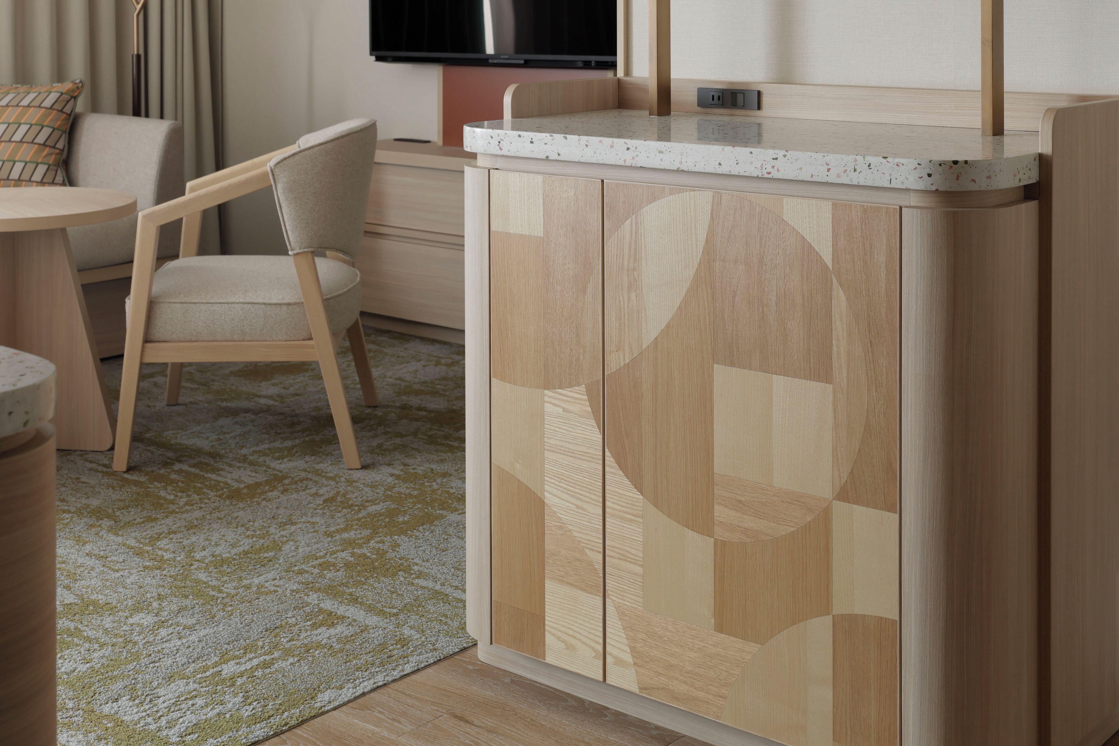 A compact hotel room with a kitchenette, a dining area with a chair, and a TV mounted on the wall.