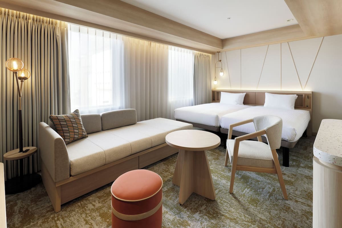 Quarto de hotel moderno com sofá, duas camas de solteiro, mesa redonda, cadeira e um pequeno banquinho.