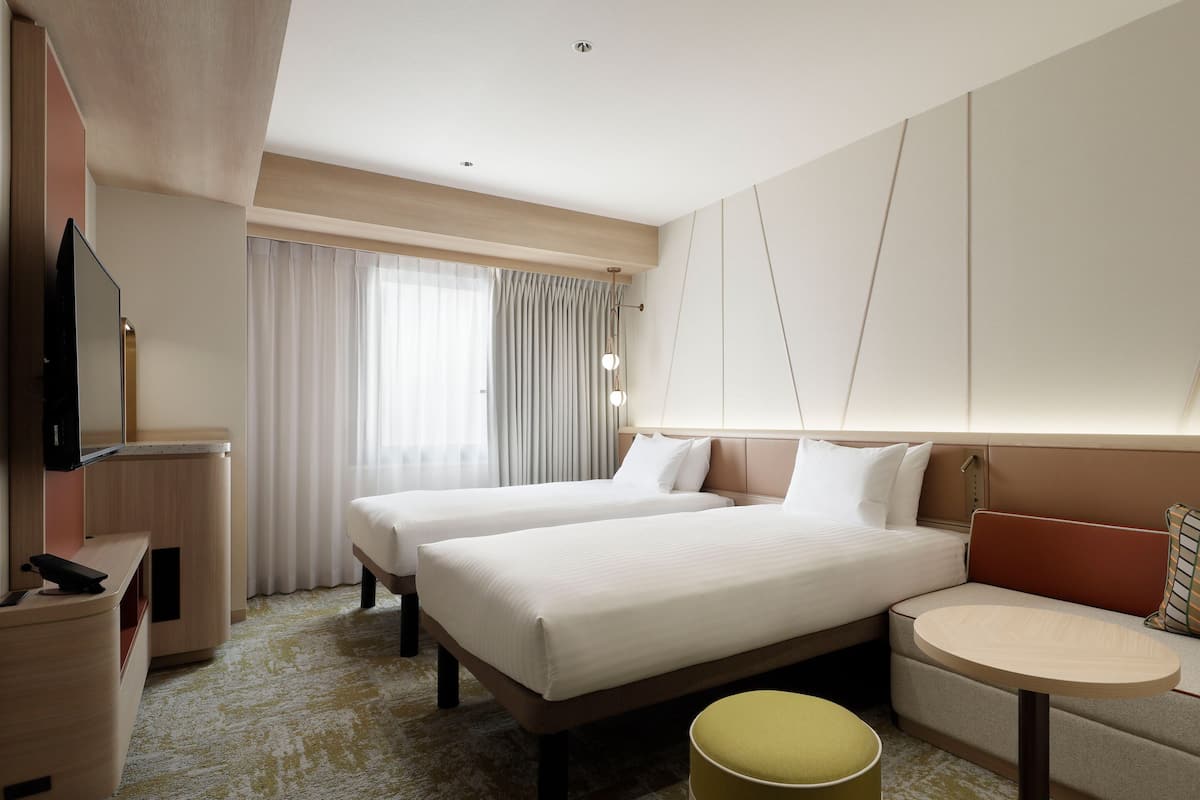 Quarto de hotel com duas camas, uma escrivaninha, uma cadeira, uma mesinha e uma televisão.