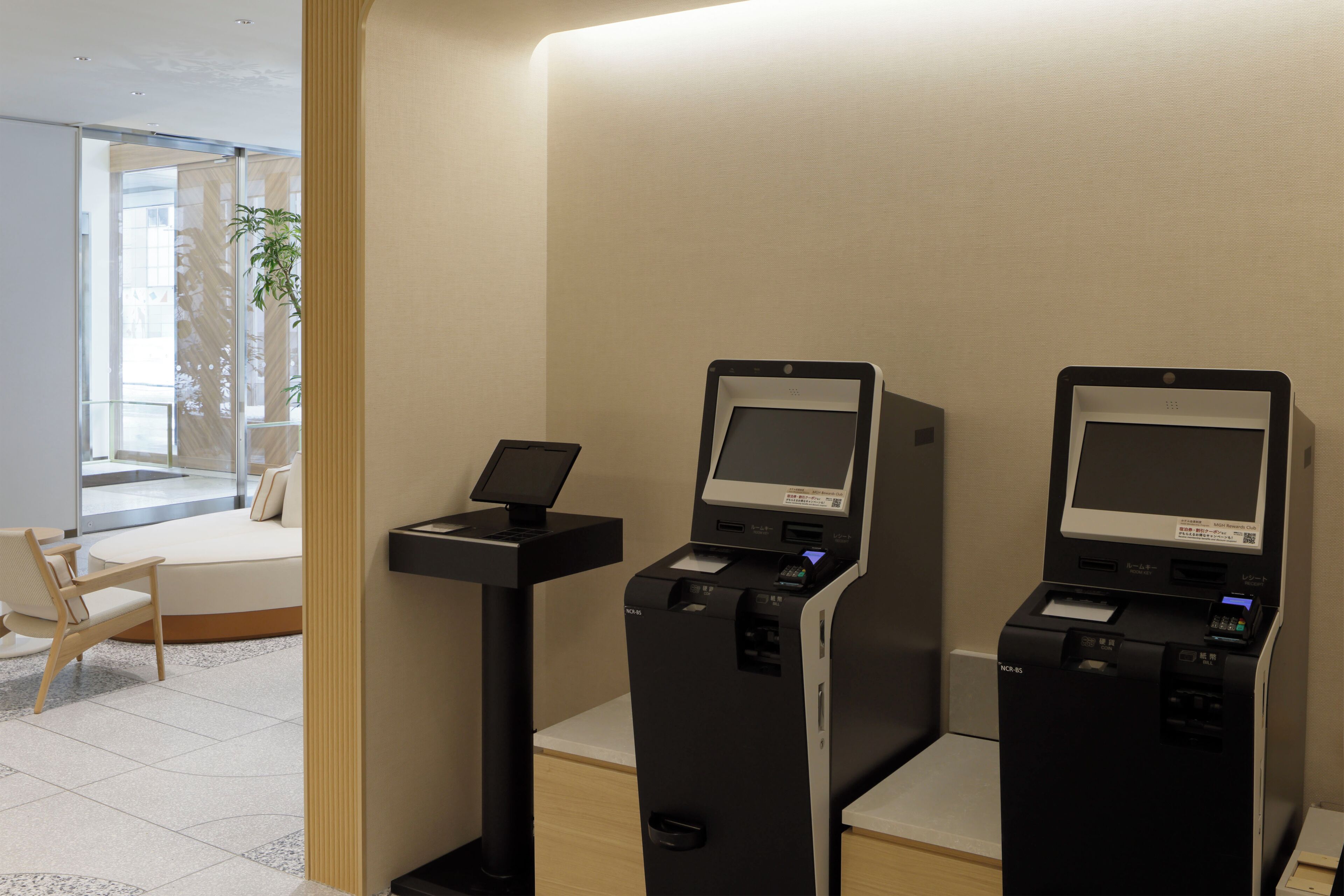 A modern hotel lobby with three self-service kiosks, a seating area with a sofa and chairs, and a large window providing natural light.