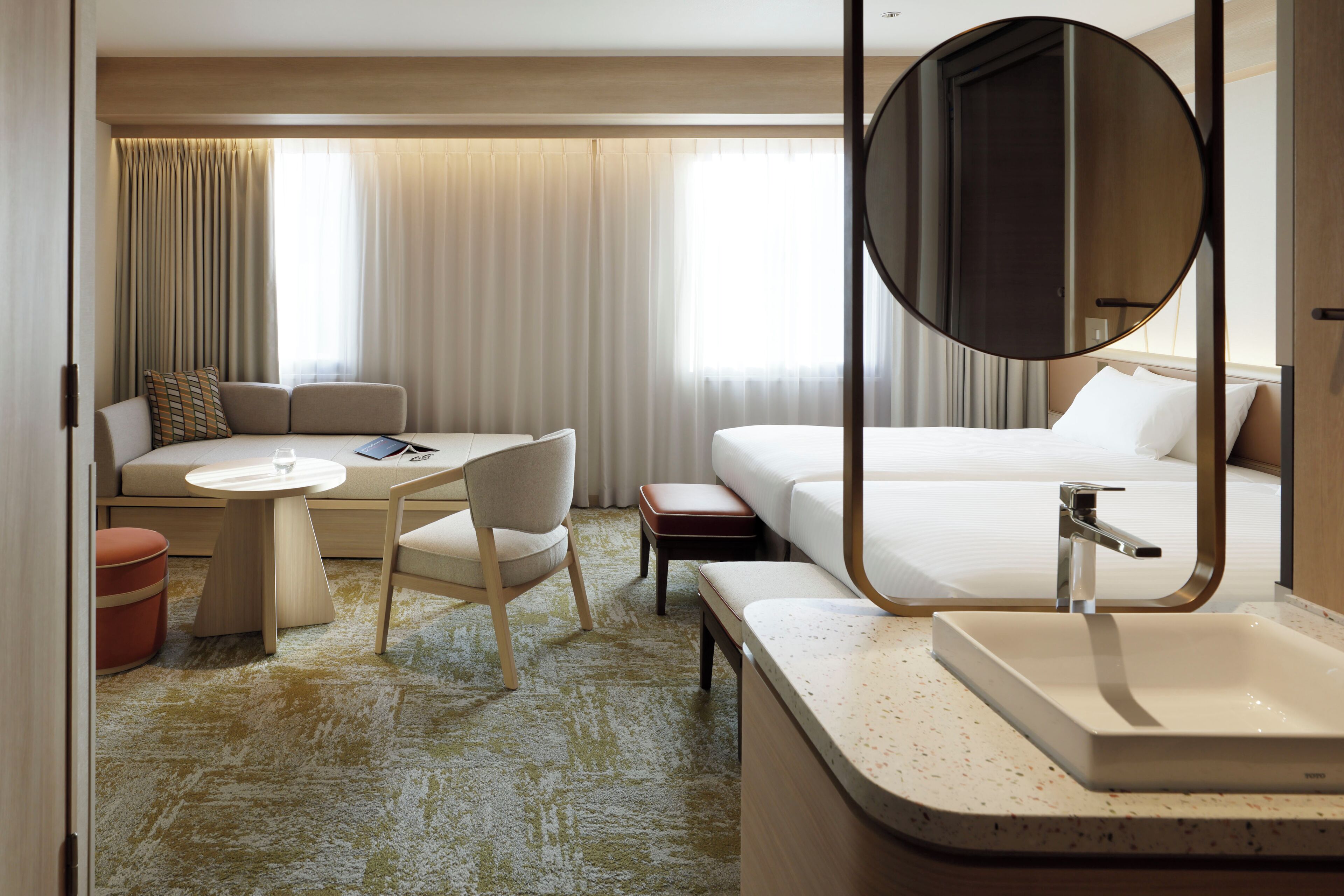 A hotel room with a bed, a round mirror, a sink, a small table, a chair, and a sofa.