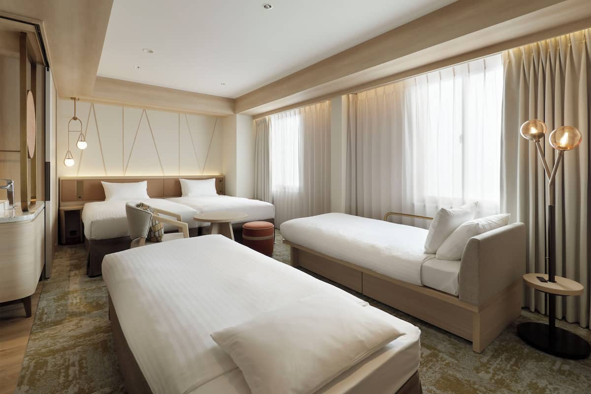 Quarto de hotel com uma cama grande, duas camas menores, uma escrivaninha e uma cadeira. Há cortinas nas janelas e um abajur.