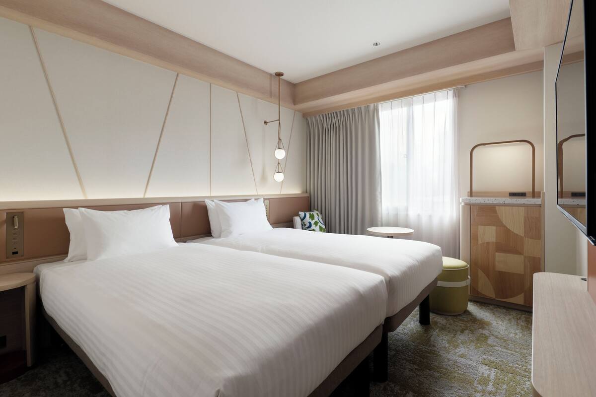 Quarto de hotel com duas camas, mesa de cabeceira, mesa redonda, espelho e janela com cortinas.