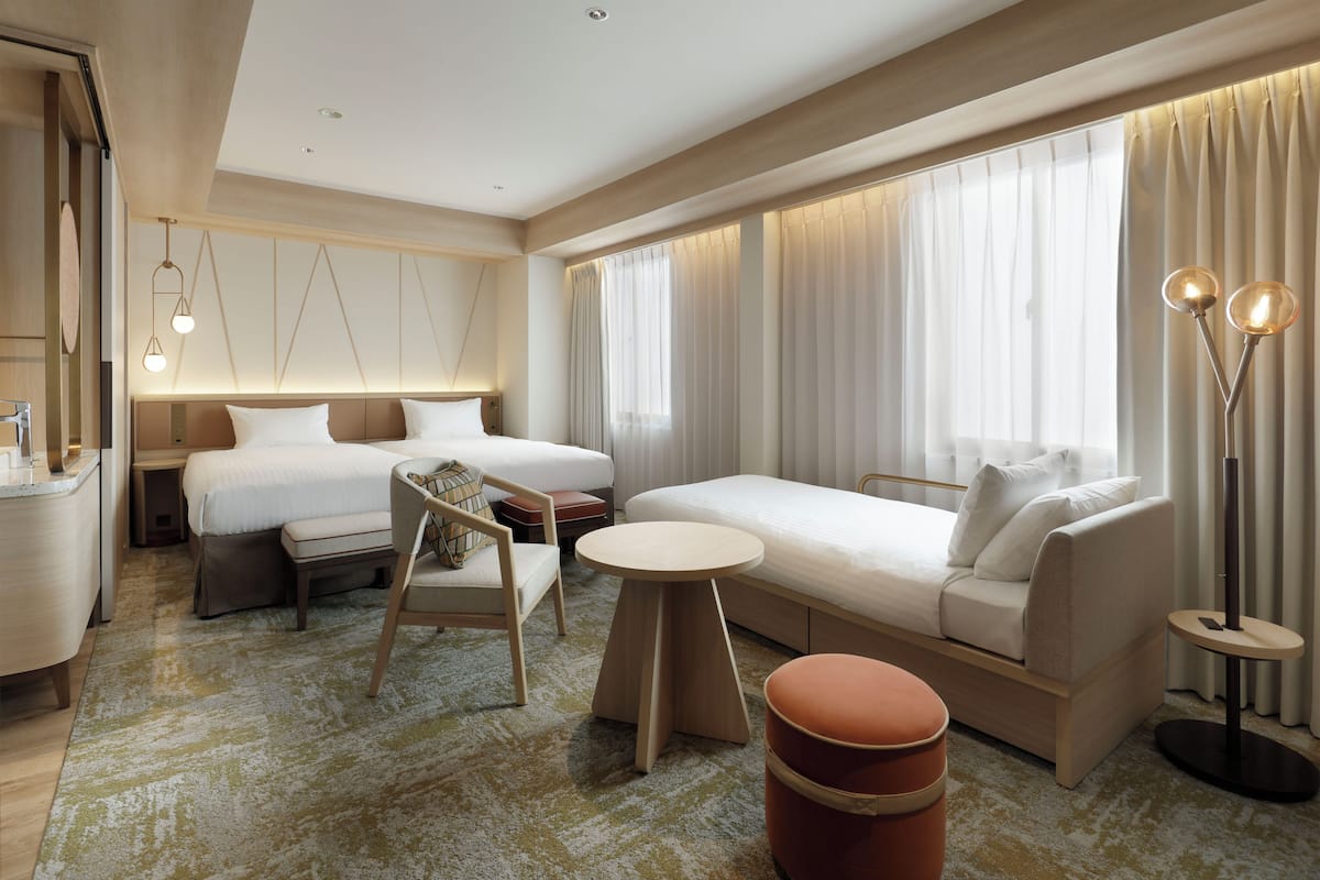 Quarto de hotel moderno com uma cama grande, um sofá, uma mesa pequena e uma cadeira. Há cortinas nas janelas e um abajur.