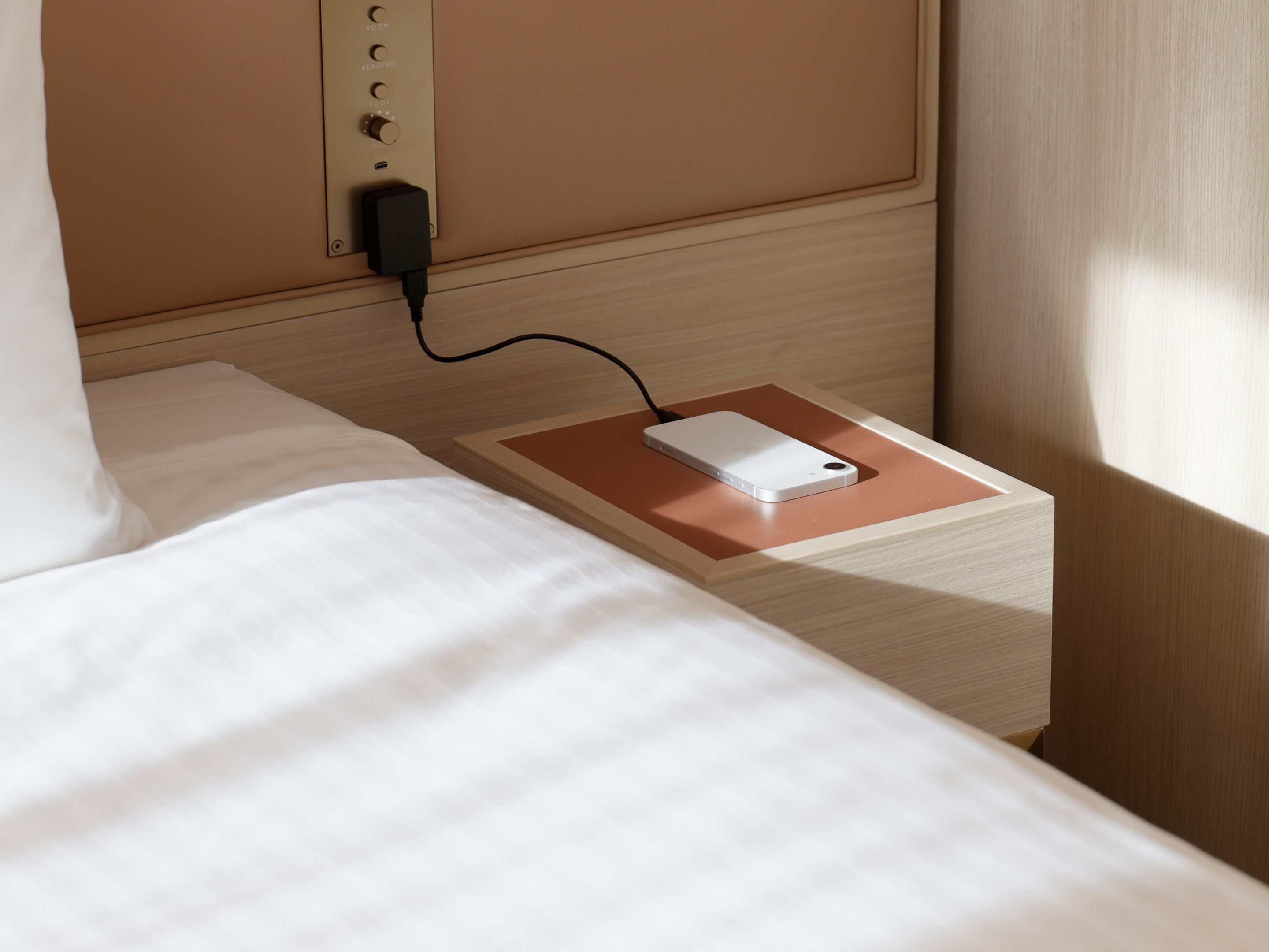 A bed with a white comforter and a wooden headboard. A white device is placed on a wooden nightstand beside the bed, connected to a power outlet on the wall.