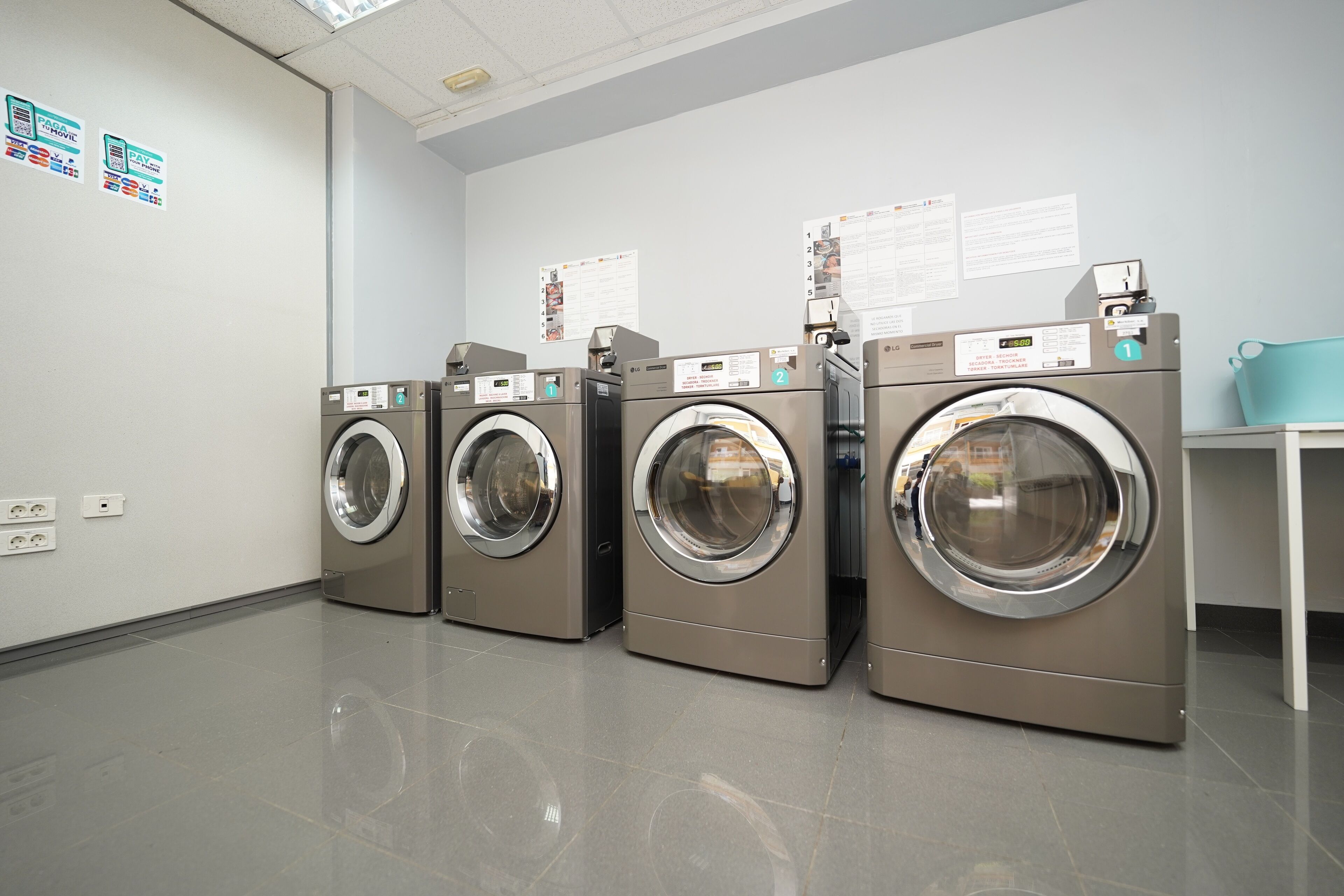 A laundry room with four front-loading washing machines, a blue trash bin, and informational posters on the wall.