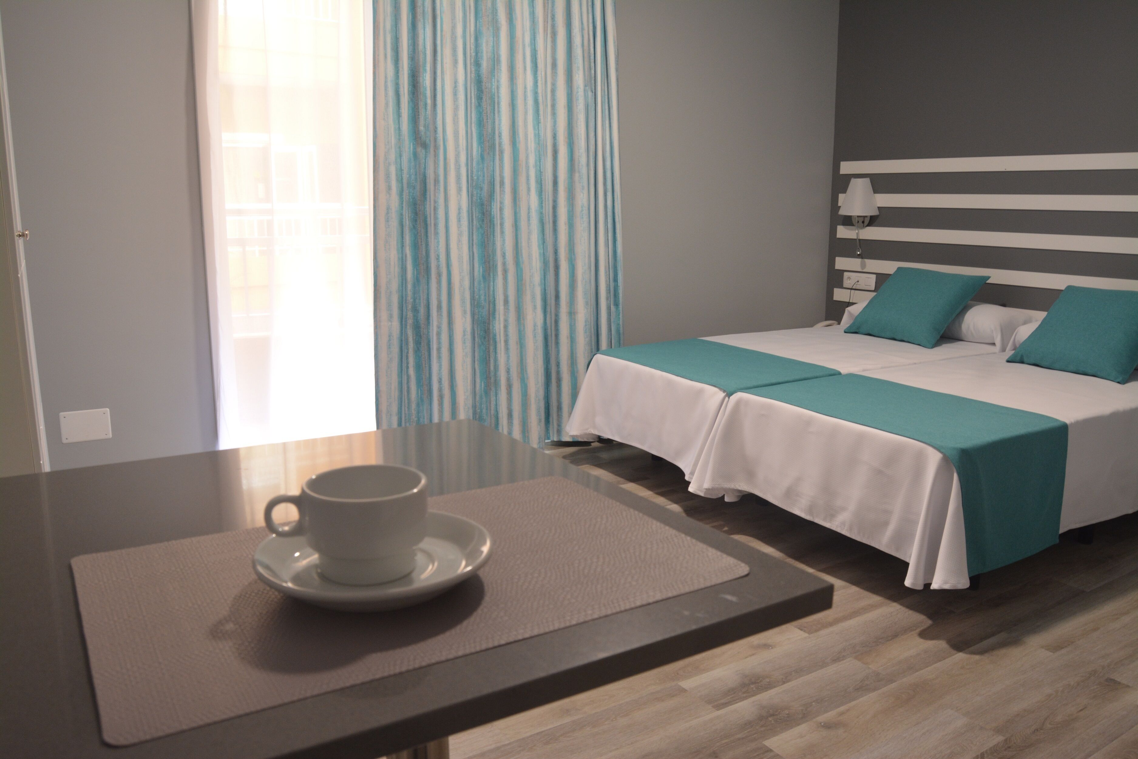 A hotel room with a bed, a bedside table with a cup and saucer, and a window with curtains.