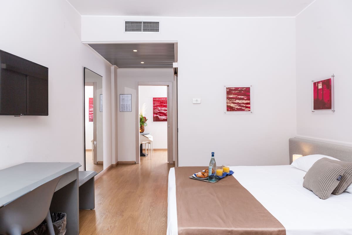 Une chambre d’hôtel moderne, équipée d’un lit, d’une table de chevet et d’un bureau avec une chaise. Les murs sont ornés d’œuvres d’art et une porte donne accès à une autre pièce.