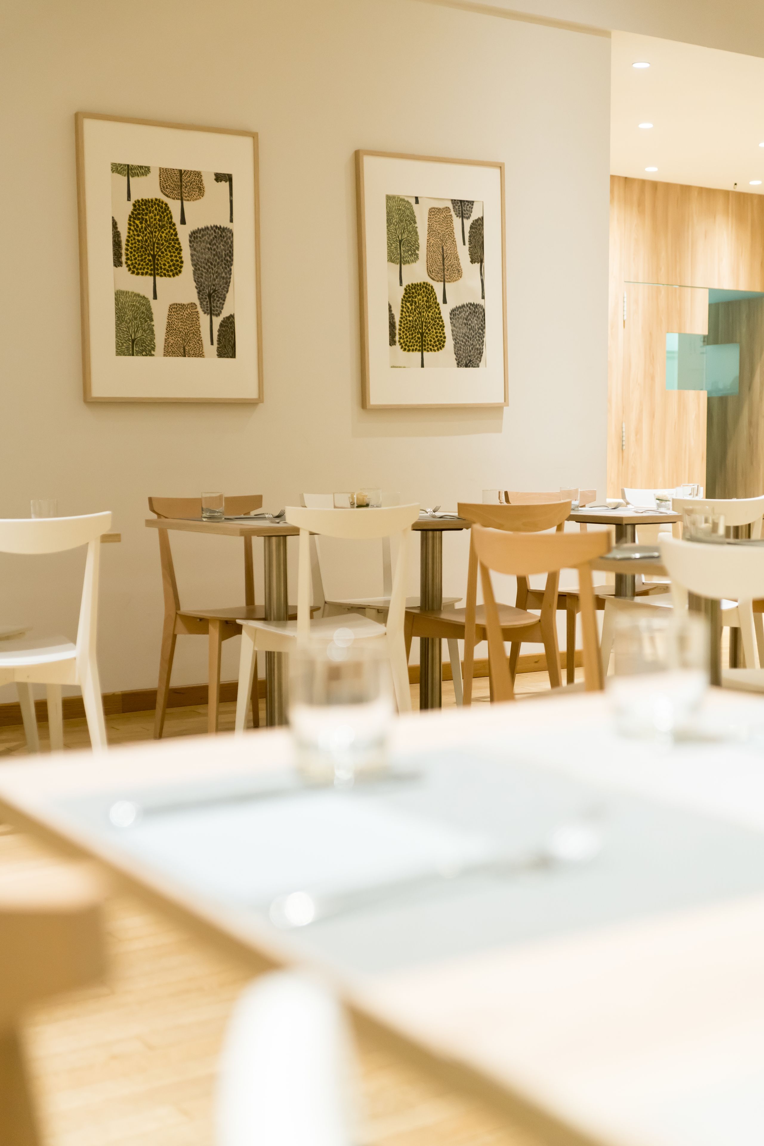A dining area with tables and chairs, framed botanical prints on the wall, and a wooden cabinet.