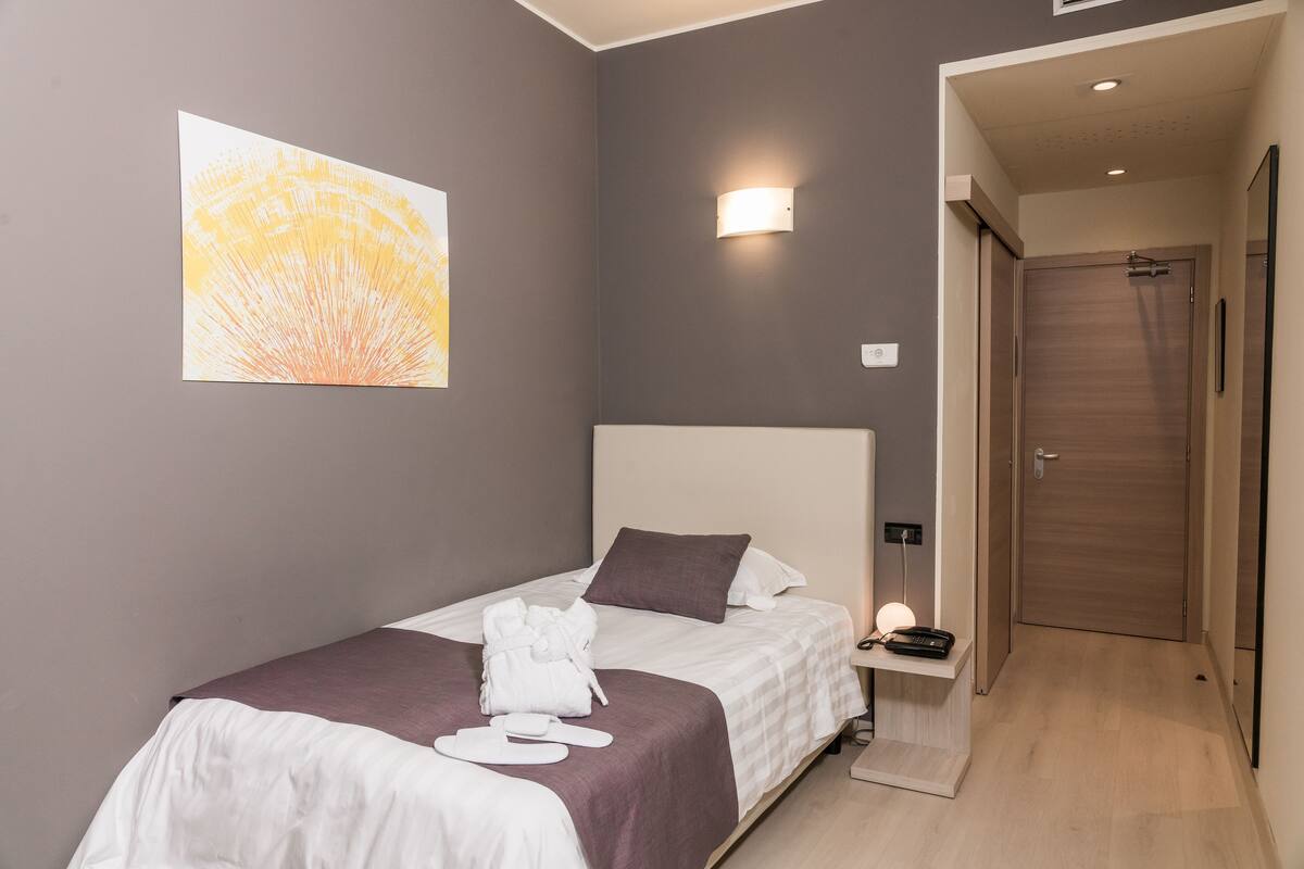 Une chambre d’hôtel moderne, équipée d’un lit simple, d’une table de chevet avec une lampe, d’un peignoir et d’un téléphone.