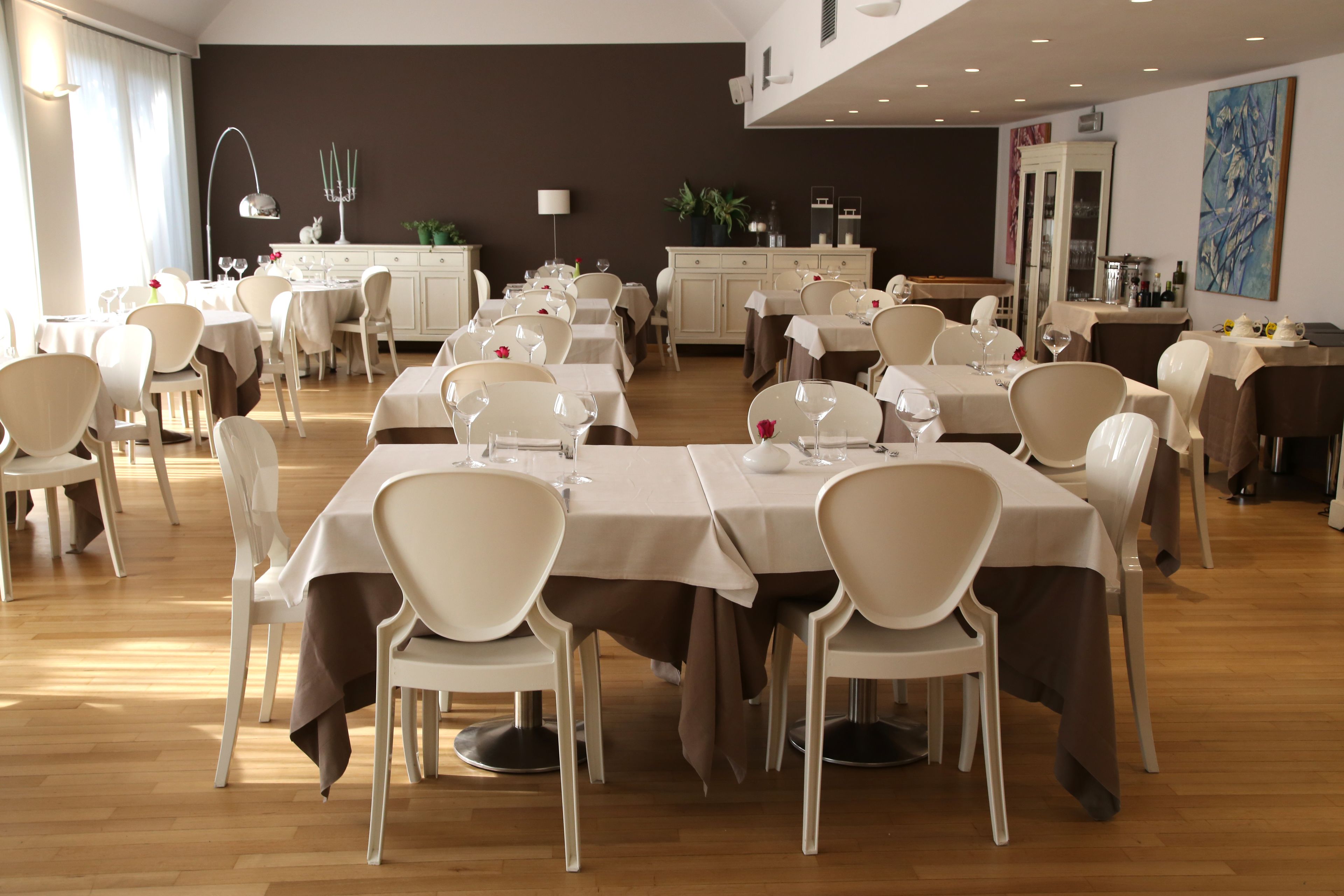 Un espace repas avec plusieurs tables et chaises, une cuisine à l’arrière-plan et un grand tableau accroché au mur.